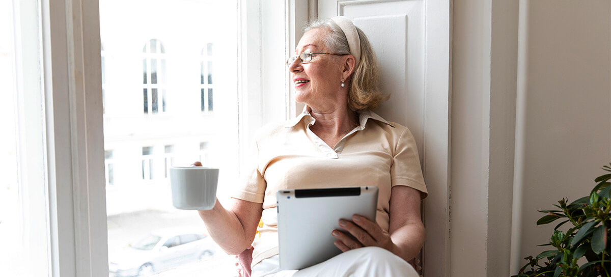 Elderly woman looking out window with coffee and iPad.